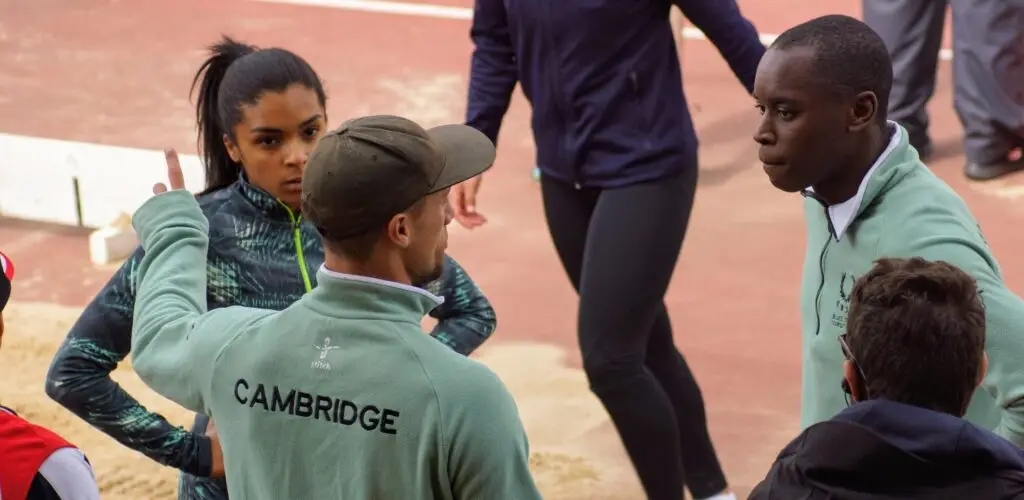 Male coaches advising female athlete on the track. Coach is wearing a light green sweatshirt with Cambridge written across the back.