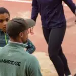 Male coaches advising female athlete on the track. Coach is wearing a light green sweatshirt with Cambridge written across the back.