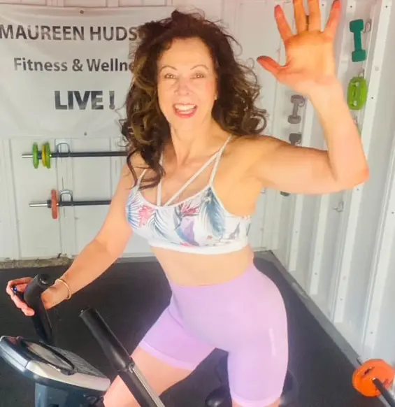 Maureen Hudson smiling and waving on a cross-fit machine
