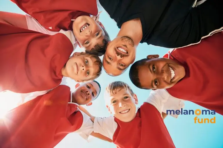 A soccer team and their coach in a huddle looking down and smiling at the ground underneath a sunny, blue sky