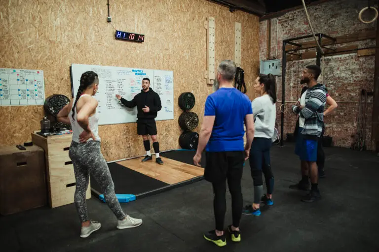 Coach at a gym gym standing in front of a whiteboard pointing at it while explaining something to a class.