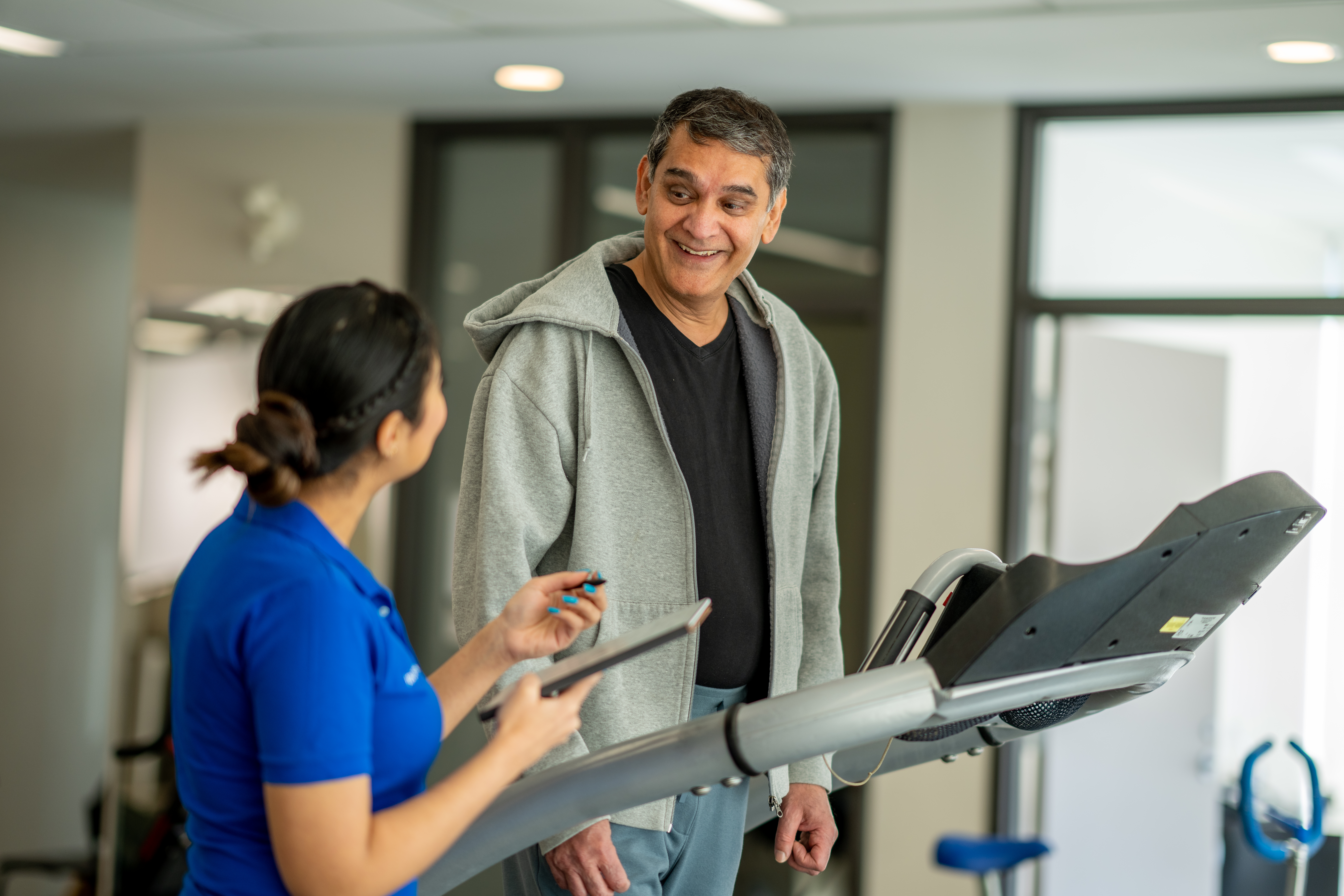 A senior gentleman walks on a treadmill in a gym during a rehabilitation appointment. His female trainer is standing beside him as she guides his exercise and takes notes.