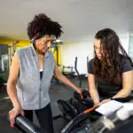 Female fitness instructor helping senior woman on treadmill in the gym
