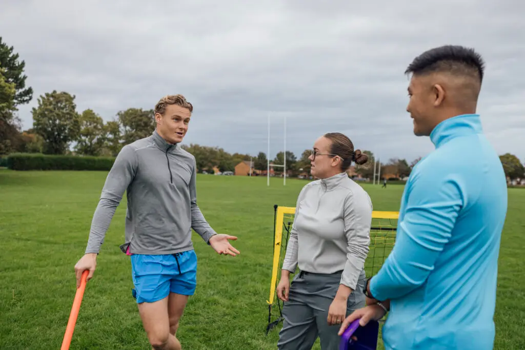 Small group of coaches standing a pitch. They are setting up equipment talking together, talking while waiting for their team.