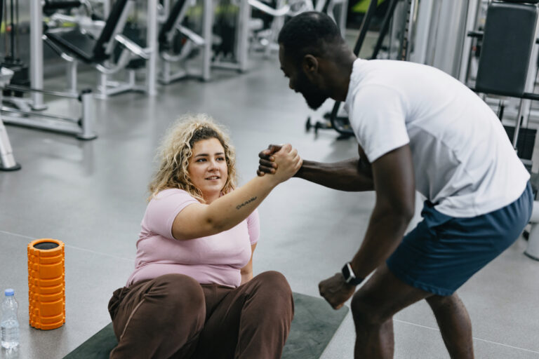 Two individuals in a gym shaking hands while one is getting up from the mat, showing support and camaraderie after their exercise session, celebrating their progress.