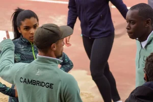 Male coaches advising female athlete on the track. Coach is wearing a light green sweatshirt with Cambridge written across the back.